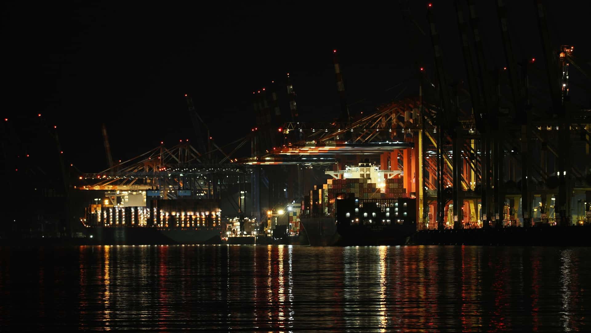 A container terminal quay viewed across the harbour at night, its cranes and stacked boxes picked out only by amber worklights, the whole scene doubled in the still water below.