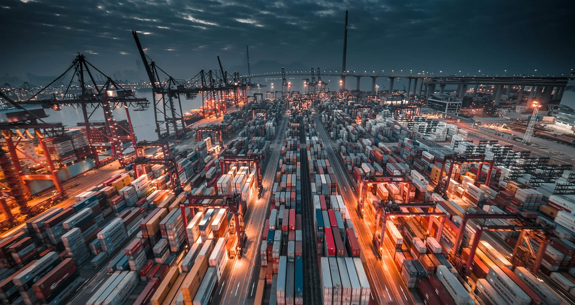 A container terminal at dusk seen from above: thousands of stacked boxes in long ranks lit orange by sodium floodlights, a line of ship-to-shore gantry cranes along the quay, and a dark storm sky over the harbour beyond.