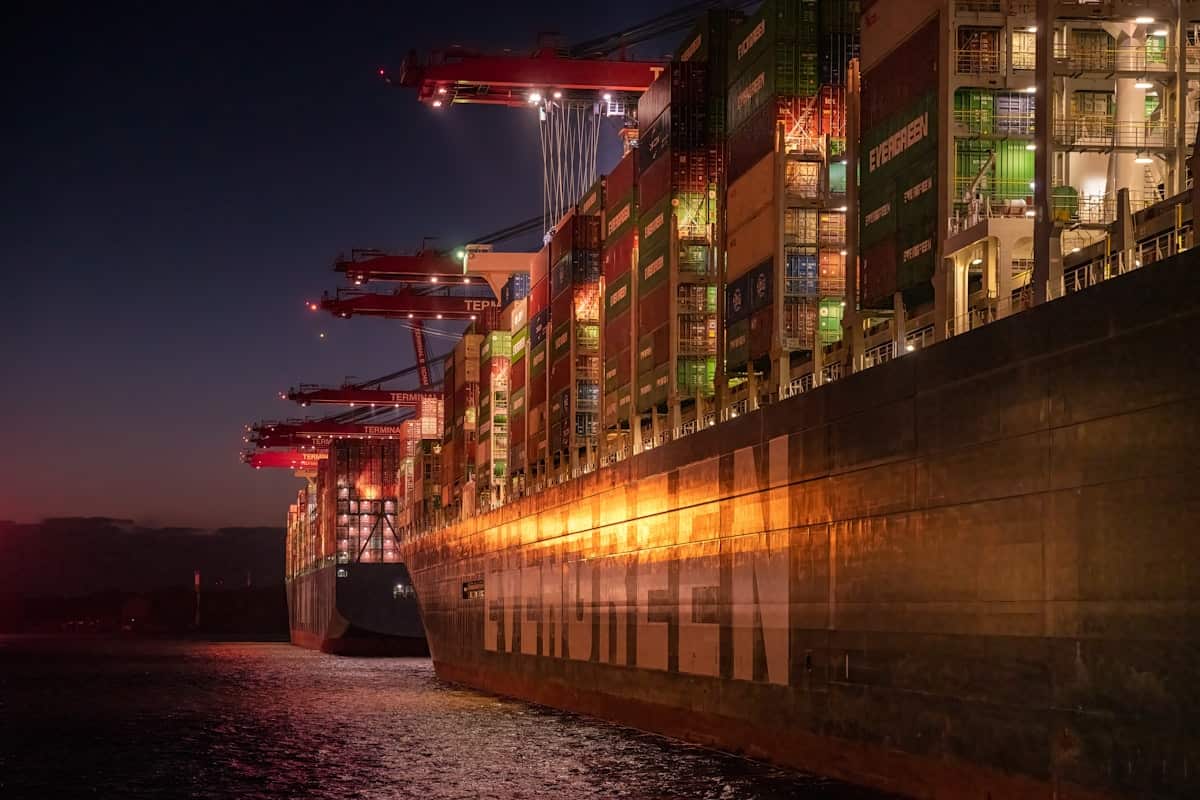 A container ship lying alongside at Waltershofer Hafen in the Port of Hamburg at dusk, its long hull glowing under crane floodlights and its deck stacked several boxes high.