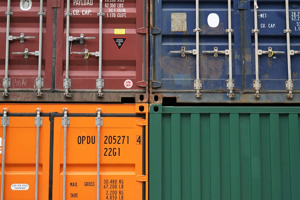 A close view of shipping container end doors in weathered corrugated steel, an orange box stencilled with its unit number and ISO type code above the payload and tare weight plate.