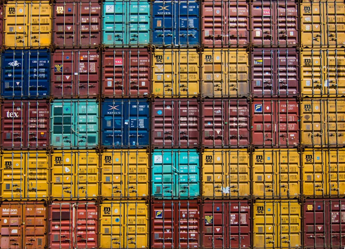A wall of shipping containers stacked end-on and filling the frame, their corrugated doors and vertical locking bars forming a grid of yellow, oxide red, teal and blue.
