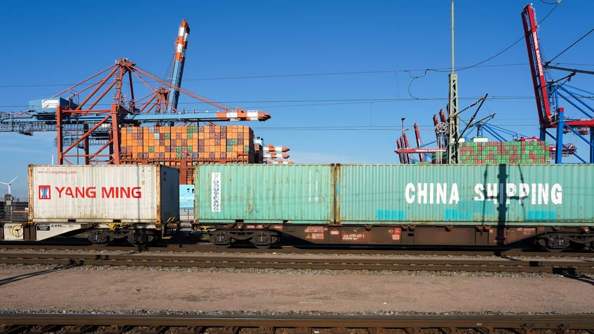 Yang Ming and China Shipping containers riding low on rail flatcars at a port intermodal yard, with red portal cranes and further stacks standing behind them.