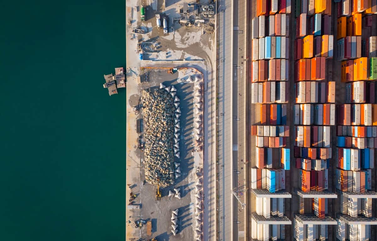 A straight-down aerial of a container yard, deep green harbour water cut hard against the concrete quay edge, with dense parallel ranks of containers filling the land side.