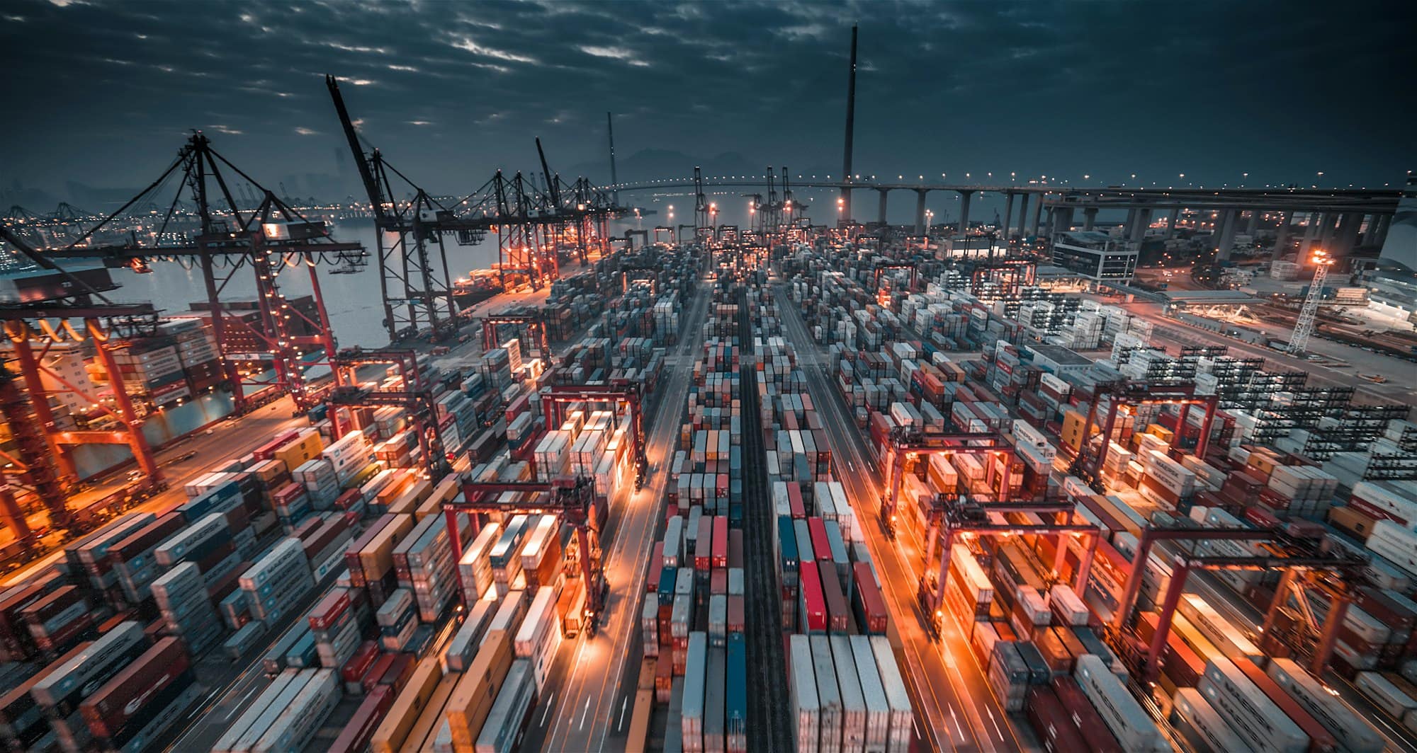 A container terminal at dusk seen from above: thousands of stacked boxes in long ranks lit orange by sodium floodlights, a line of ship-to-shore gantry cranes along the quay, and a dark storm sky over the harbour beyond.